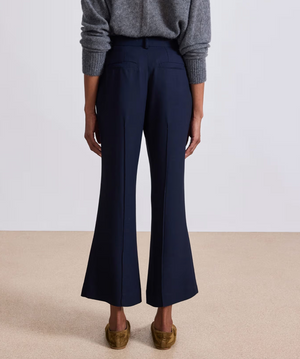 Shown from the back, a person wears the Apiece Apart Rene Flare Trouser in navy with a grey sweater and brown loafers, standing on a beige floor against a plain background.