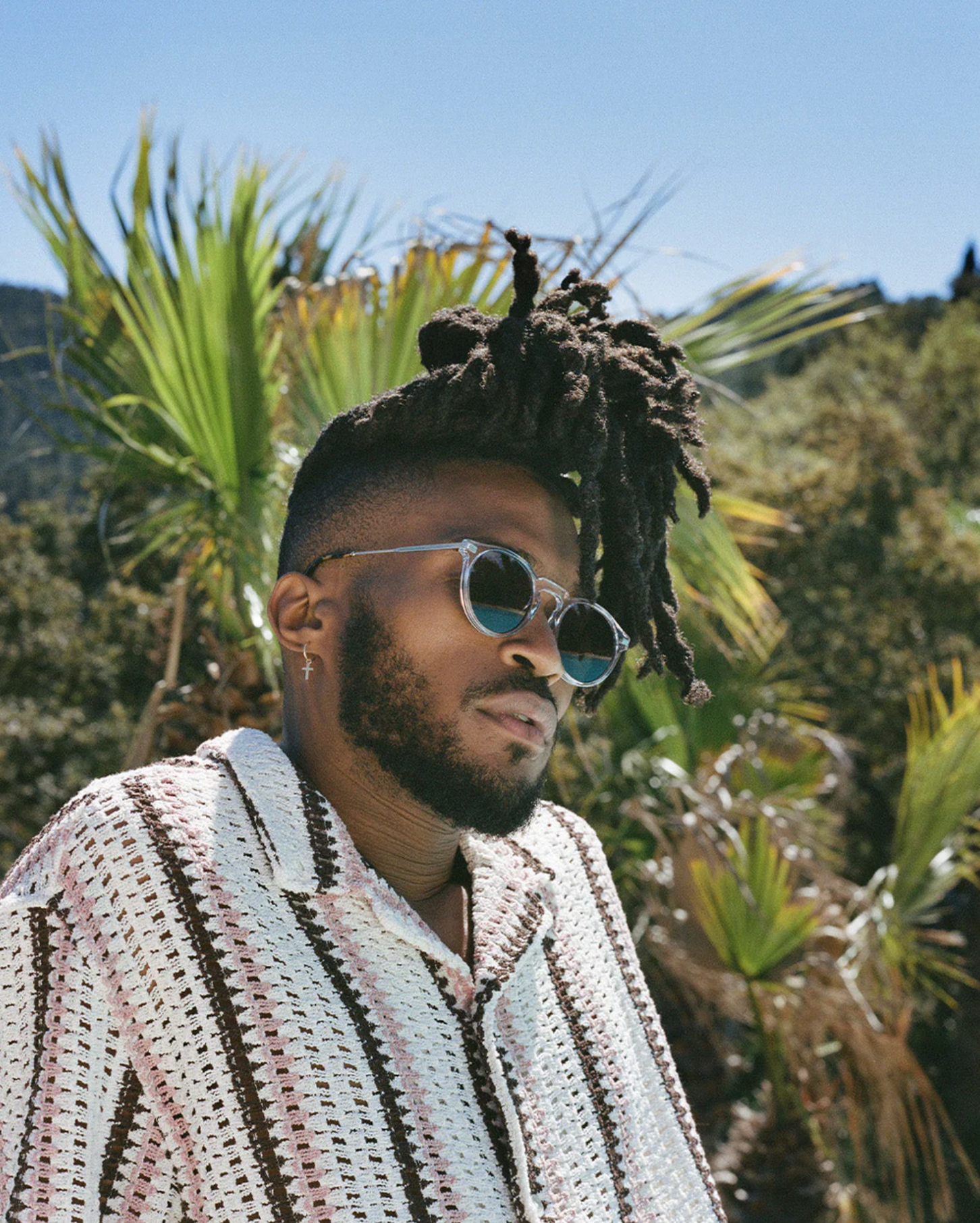 A man wearing Jimmy Fairly's The Noto sunglasses, which have a lightweight acetate frame, stands outdoors in a patterned shirt amid palm trees and greenery under a clear blue sky.
