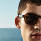 Close-up of a man with short hair wearing Jimmy Fairly's The Ezra bio-acetate tortoiseshell sunglasses, set against a blurred ocean background under a clear sky.