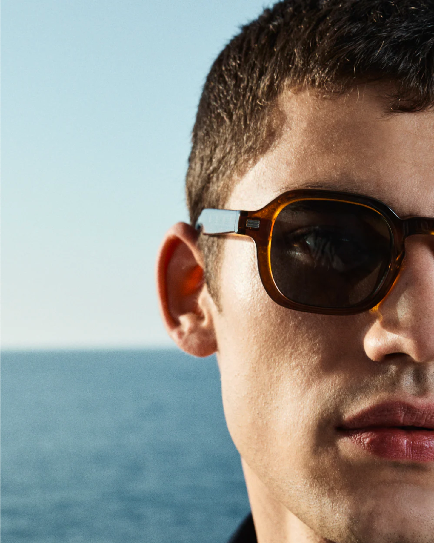 Close-up of a man with short hair wearing Jimmy Fairly's The Ezra bio-acetate tortoiseshell sunglasses, set against a blurred ocean background under a clear sky.