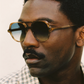 A man with short curly hair, a mustache, and wearing Jimmy Fairly's The Lars brown cellulose acetate sunglasses and a plaid shirt looks to the side.