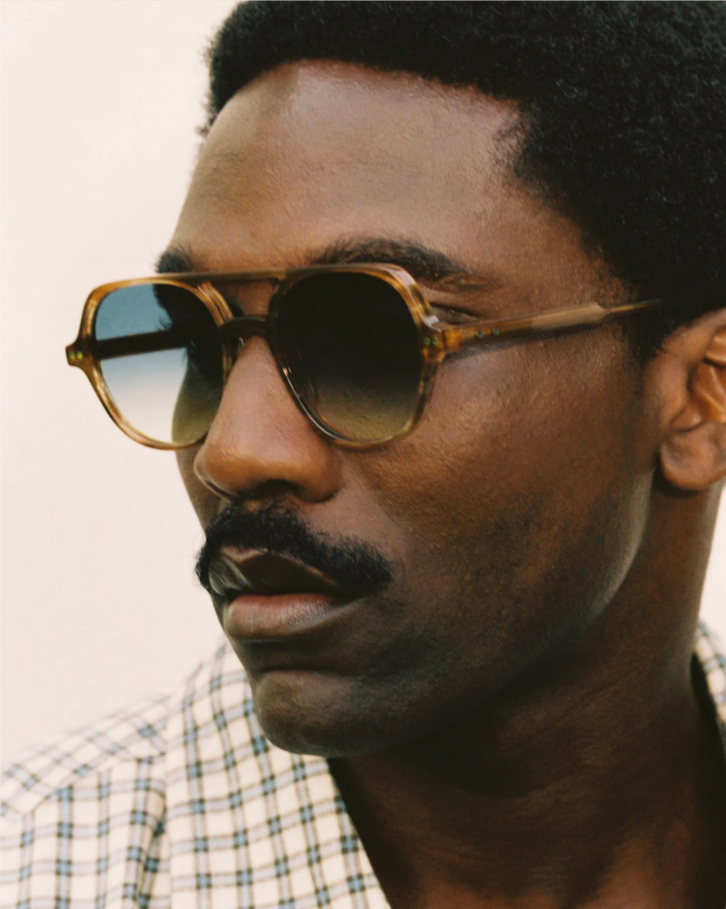 A man with short curly hair, a mustache, and wearing Jimmy Fairly's The Lars brown cellulose acetate sunglasses and a plaid shirt looks to the side.