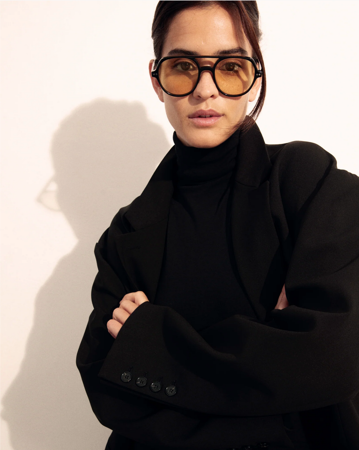 Model wearing The Lars oversized yellow-tinted sunglasses by Jimmy Fairly, featuring a bold cellulose acetate frame, paired with a black turtleneck and blazer, standing against a light background with arms crossed.