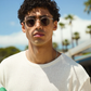 A man with curly hair wearing The Hometown round sunglasses by Jimmy Fairly, featuring a sleek cellulose acetate frame and a light textured sweater, stands outdoors with palm trees and blue sky in the background.