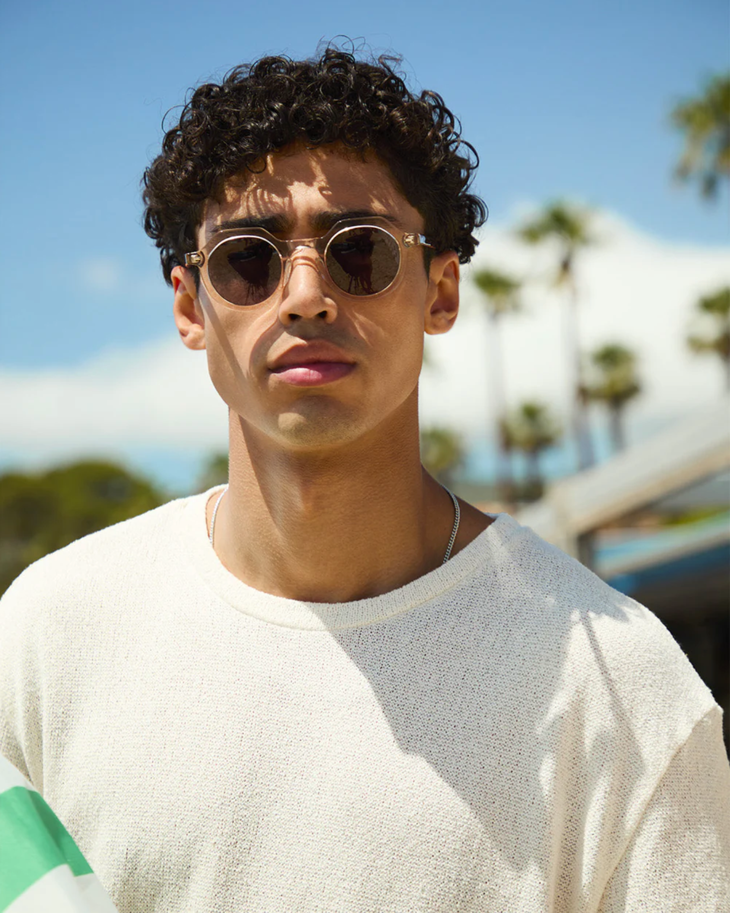 A man with curly hair wearing The Hometown round sunglasses by Jimmy Fairly, featuring a sleek cellulose acetate frame and a light textured sweater, stands outdoors with palm trees and blue sky in the background.
