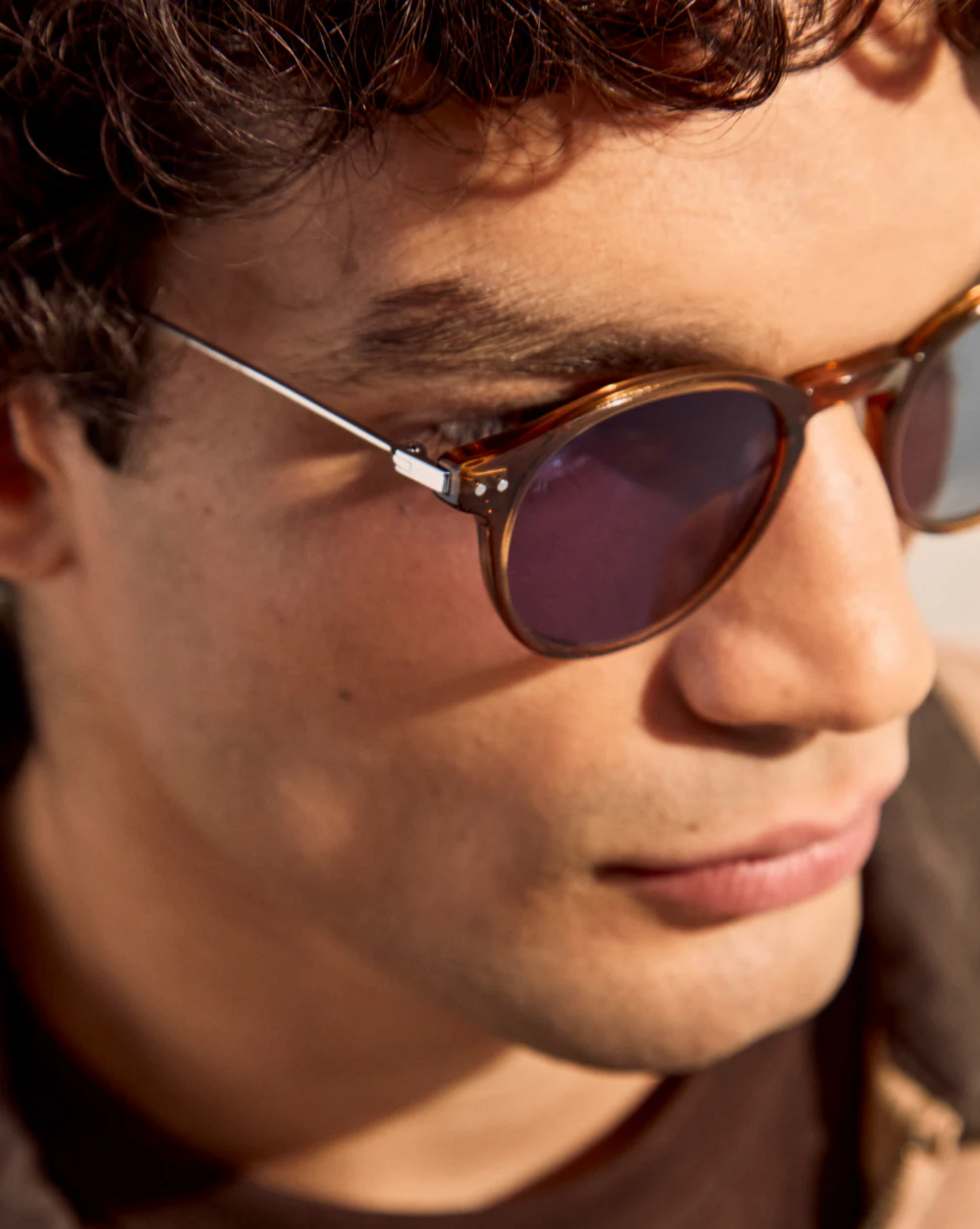 Close-up of a person with curly hair wearing The Emilio sunglasses by Jimmy Fairly, featuring brown acetate frames and looking slightly downward.