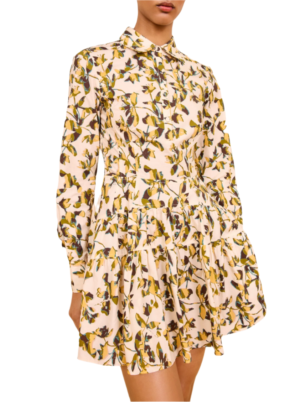 A woman wears the Ulla Johnson Nanette Mini Dress—a long-sleeve, collared poplin dress with yellow and green graphic blooms on a light background.