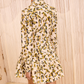 A person stands with their back to the camera, wearing the Ulla Johnson Nanette Mini Dress featuring graphic blooms and a ruffled skirt, against a beige textured background.
