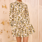 A person wearing the Ulla Johnson Nanette Mini Dress, a long-sleeve, collared poplin dress with yellow and green graphic blooms, stands indoors against a textured cream background.