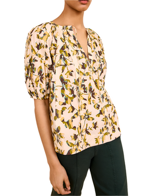 Someone is wearing the Ulla Johnson Loli Top, a light-colored, short-sleeve blouse with a multicolor abstract floral print, paired with dark pants and photographed from the waist up.