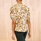 A person stands with their back to the camera wearing the Ulla Johnson Loli Top, a light floral blouse with puffed sleeves, and dark green pants against a neutral textured background.