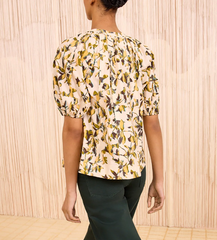 A person stands with their back to the camera wearing the Ulla Johnson Loli Top, a light floral blouse with puffed sleeves, and dark green pants against a neutral textured background.