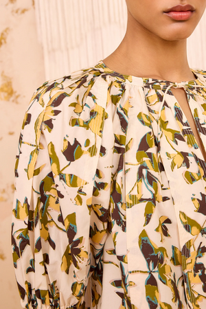 Close-up of a person wearing the Loli Top by Ulla Johnson, a cream-colored cotton poplin blouse featuring yellow, green, and brown abstract floral prints, gathered neckline details, and subtle puff sleeves.