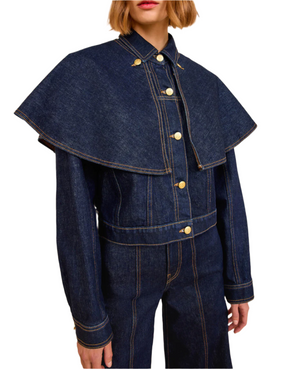 A person wearing the Ulla Johnson Agata Denim Cape Jacket, a vintage-inspired dark denim piece with a wide cape-like collar, paired with matching denim pants, shown from mid-torso up.