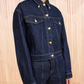 A person wears a Ulla Johnson Agata Denim Cape Jacket and matching high-waisted denim pants, standing before a beige textured background.