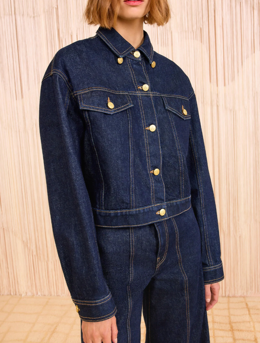 A person wears a Ulla Johnson Agata Denim Cape Jacket and matching high-waisted denim pants, standing before a beige textured background.