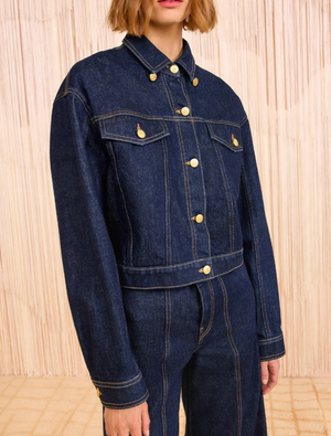 A person wears a Ulla Johnson Agata Denim Cape Jacket and matching high-waisted denim pants, standing before a beige textured background.