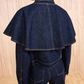 A person stands facing away, wearing the Ulla Johnson Agata Denim Cape Jacket with a wide cape collar and matching denim pants against a light background.