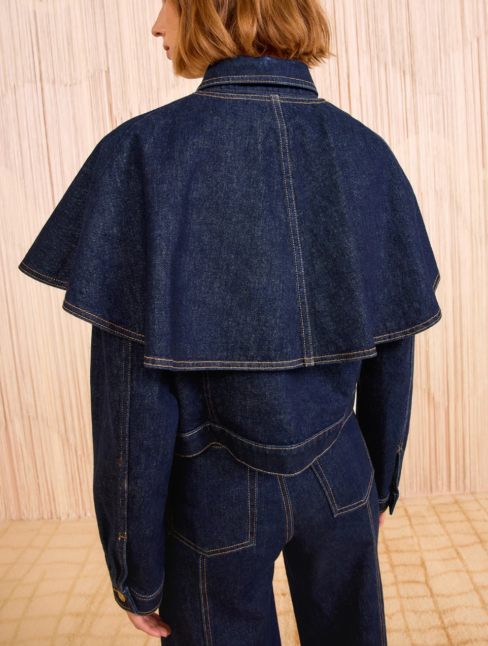 A person stands facing away, wearing the Ulla Johnson Agata Denim Cape Jacket with a wide cape collar and matching denim pants against a light background.