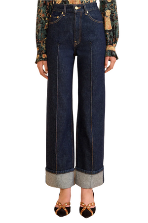 A person wears the Ulla Johnson Genevieve High Rise Jean, made from premium vintage indigo denim, styled with a patterned blouse and pointed-toe shoes featuring gold accents.