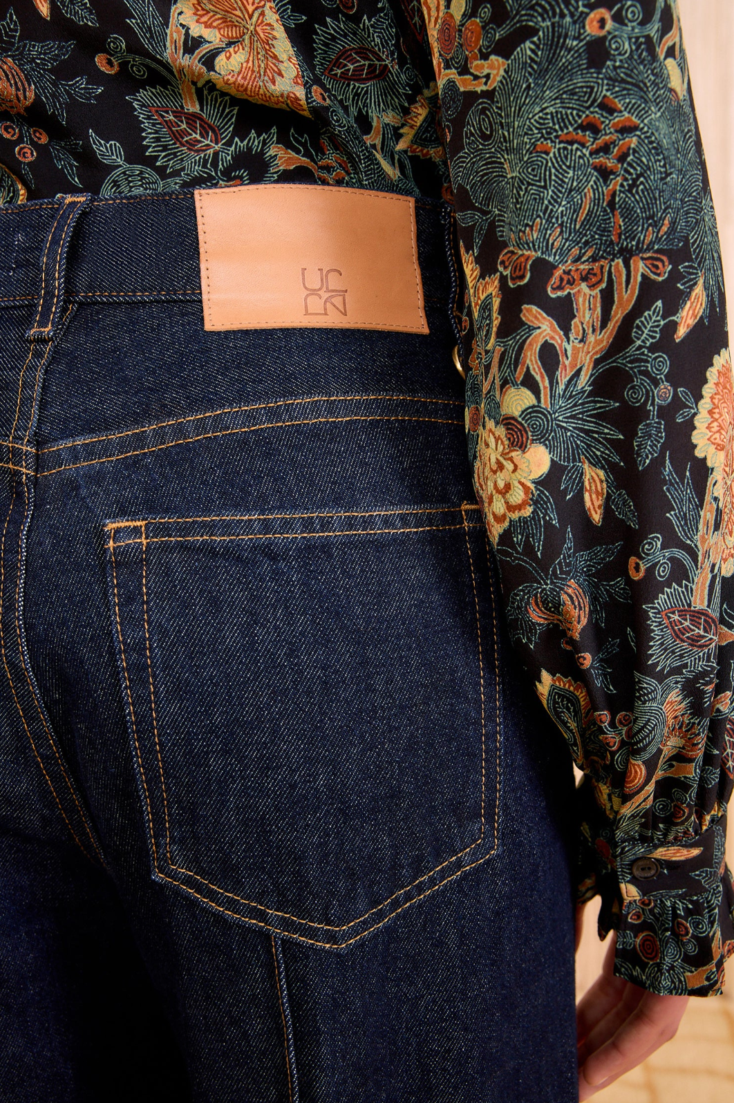 Wearing Ulla Johnson’s Genevieve High Rise Jean in vintage-inspired indigo with a tan patch, paired with a floral patterned shirt.
