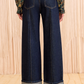 A person wears Ulla Johnson’s Genevieve High Rise Jean—vintage-inspired indigo denim with wide legs, large cuffs, and back pockets—paired with a floral long-sleeve top and black heeled sandals on a light floor.