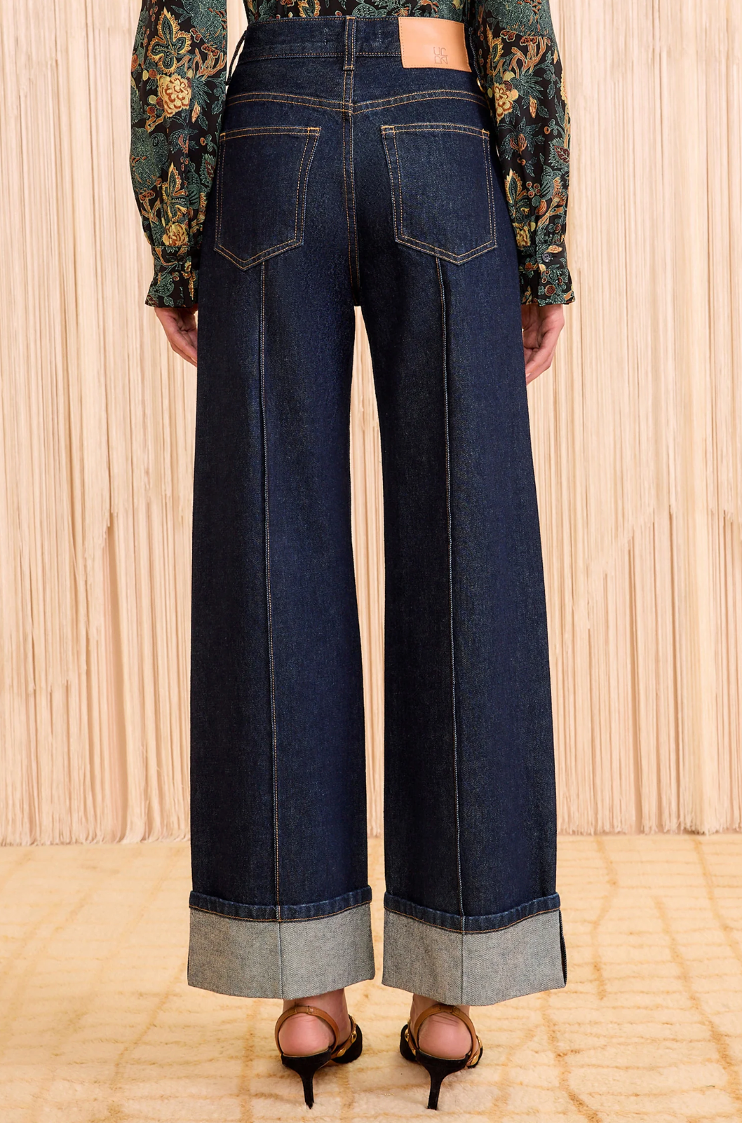 A person wears Ulla Johnson’s Genevieve High Rise Jean—vintage-inspired indigo denim with wide legs, large cuffs, and back pockets—paired with a floral long-sleeve top and black heeled sandals on a light floor.