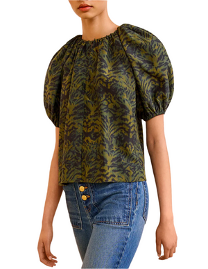 A person wearing the Ulla Johnson Talie Silk Blouse in green and navy ikat print with puffed sleeves, paired with high-waisted blue jeans featuring gold buttons.