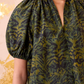 A woman wears the Ulla Johnson Talie Silk Blouse, featuring green and black ikat print and a split neckline, paired with blue jeans. The light background has gold accents.