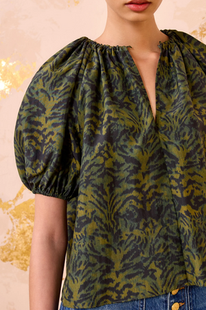 A woman wears the Ulla Johnson Talie Silk Blouse, featuring green and black ikat print and a split neckline, paired with blue jeans. The light background has gold accents.