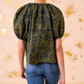 A model wears the Ulla Johnson Talie Silk Blouse in green and black ikat print with puffed sleeves, paired with blue jeans, standing against a light background with gold accents.