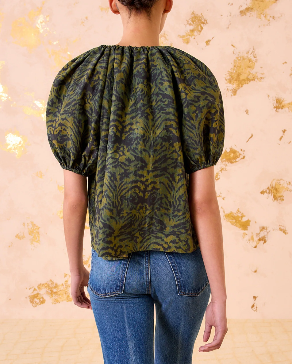 A model wears the Ulla Johnson Talie Silk Blouse in green and black ikat print with puffed sleeves, paired with blue jeans, standing against a light background with gold accents.