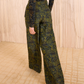 A person wears the Ulla Johnson Charlie Silk Pant in dark green ikat animal print, paired with black heeled sandals, standing on a beige carpet before a wooden backdrop.