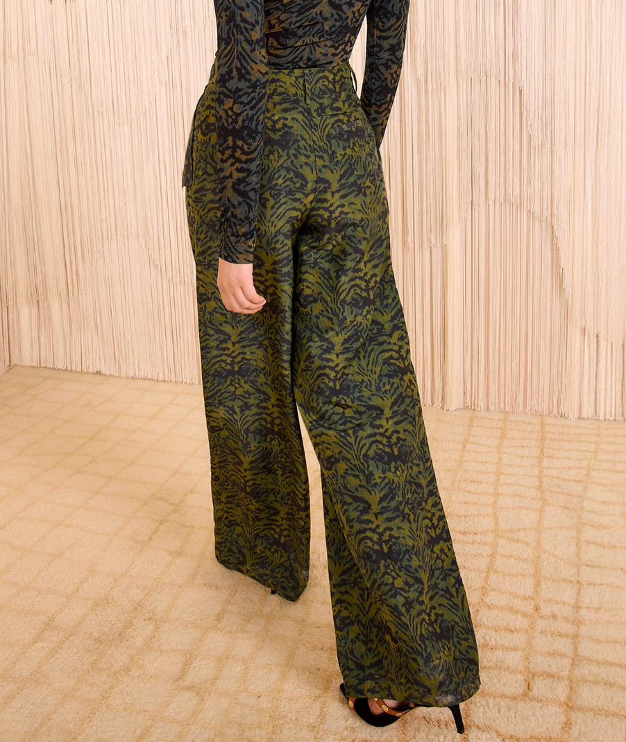 A person wears the Ulla Johnson Charlie Silk Pant in dark green ikat animal print, paired with black heeled sandals, standing on a beige carpet before a wooden backdrop.