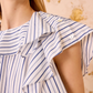 A close-up of a woman wearing the Ulla Johnson Amaia Top, a blue striped blouse with ruffled shoulder details made from soft cotton shirting, set against a beige and gold textured background.