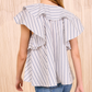 A person wearing the Ulla Johnson Amaia Top, a blue striped blouse with ruffled sleeves, pairs it with blue jeans and stands facing a beige textured wall.