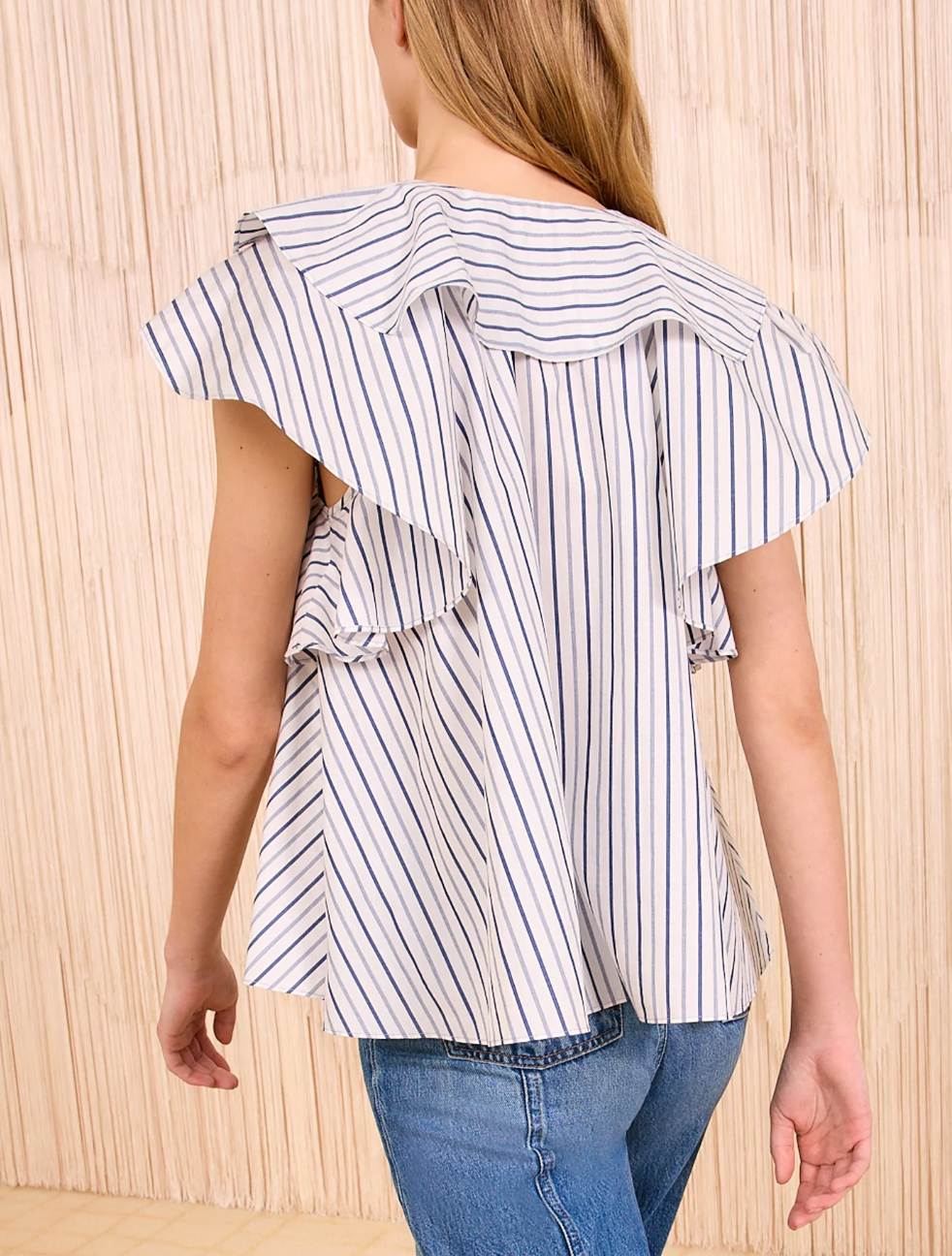 A person wearing the Ulla Johnson Amaia Top, a blue striped blouse with ruffled sleeves, pairs it with blue jeans and stands facing a beige textured wall.