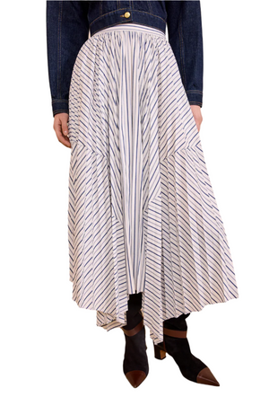Wearing the Ulla Johnson Yasna Midi Skirt, a white cotton piece with diagonal blue stripes and an asymmetric hem, paired with a dark denim jacket and black pointed-toe shoes with brown heels.
