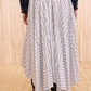 A person wears the Ulla Johnson Yasna Midi Skirt—white cotton with blue stripes and an asymmetric hem—paired with a dark denim jacket and black ankle boots, standing indoors on a light patterned floor.