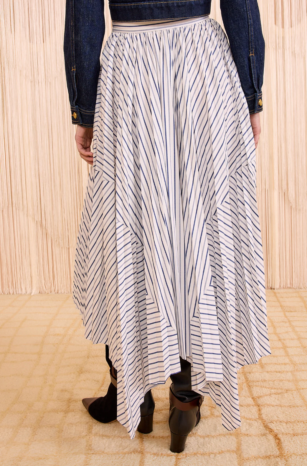 A person wears the Ulla Johnson Yasna Midi Skirt—white cotton with blue stripes and an asymmetric hem—paired with a dark denim jacket and black ankle boots, standing indoors on a light patterned floor.