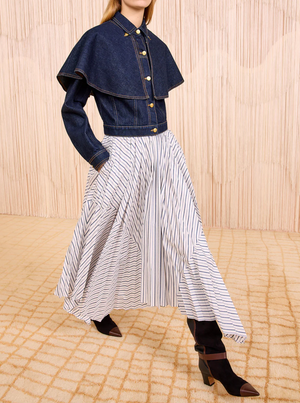 Person wearing a structured dark denim jacket with wide shoulder flaps, paired with the Ulla Johnson Yasna Midi Skirt—a flowy cotton piece featuring an asymmetric hem and thin blue stripes—and black pointed-toe boots.