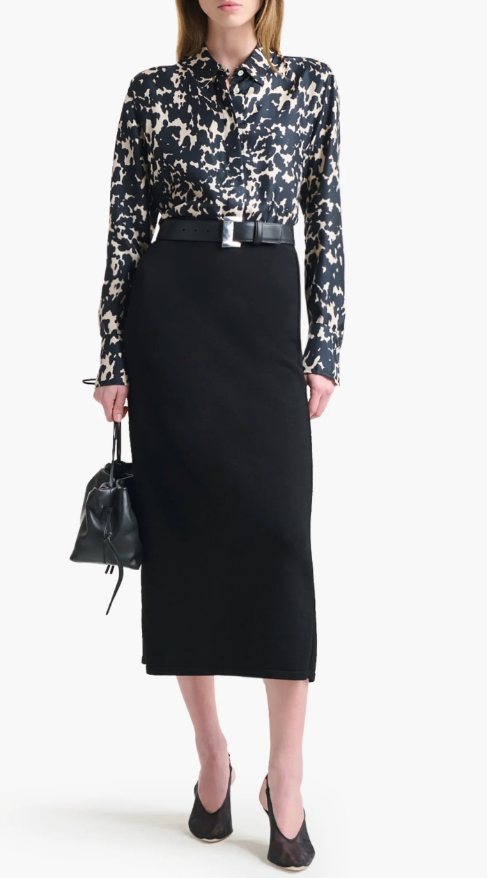 A person wears the Altuzarra Chika Top—a black and white patterned silk shirt with a concealed button front—styled with a black belt, midi skirt, heels, and a small black handbag for a relaxed look.