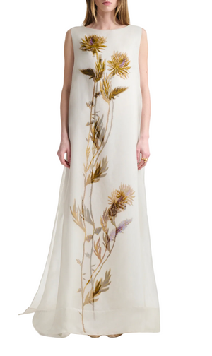 A woman wears the Altuzarra Jools Dress, a sleeveless, floor-length white silk gown featuring a vertical botanical print with large yellow and purple thistle flowers and green stems.