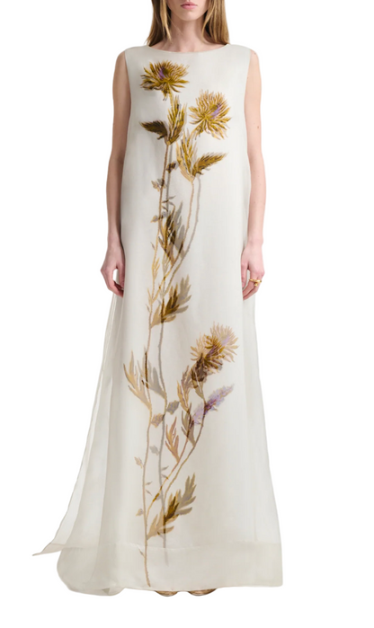 A woman wears the Altuzarra Jools Dress, a sleeveless, floor-length white silk gown featuring a vertical botanical print with large yellow and purple thistle flowers and green stems.