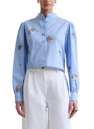 A person wears the Altuzarra Margot Top, a blue and white vertical striped cotton-poplin shirt with floral patterns and front buttons, paired with high-waisted white pants.