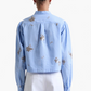 A person with dark hair in a bun is shown from the back wearing the Altuzarra Margot Top— a modern blue and white vertically striped cotton-poplin shirt with floral patterns—paired with white pants.