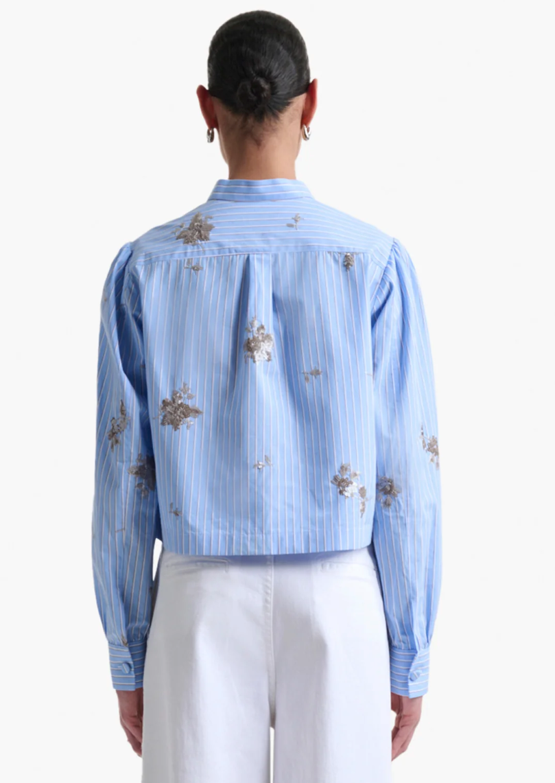 A person with dark hair in a bun is shown from the back wearing the Altuzarra Margot Top— a modern blue and white vertically striped cotton-poplin shirt with floral patterns—paired with white pants.