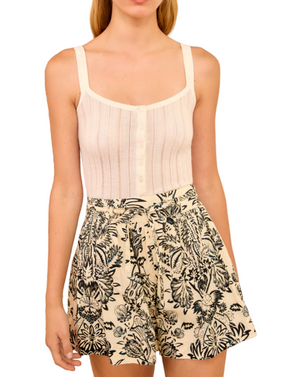 A woman wears a sleeveless, buttoned white ribbed top and Ulla Johnson Rowan Belted Shorts, which are high-waisted ivory poplin featuring a black floral leaf print.