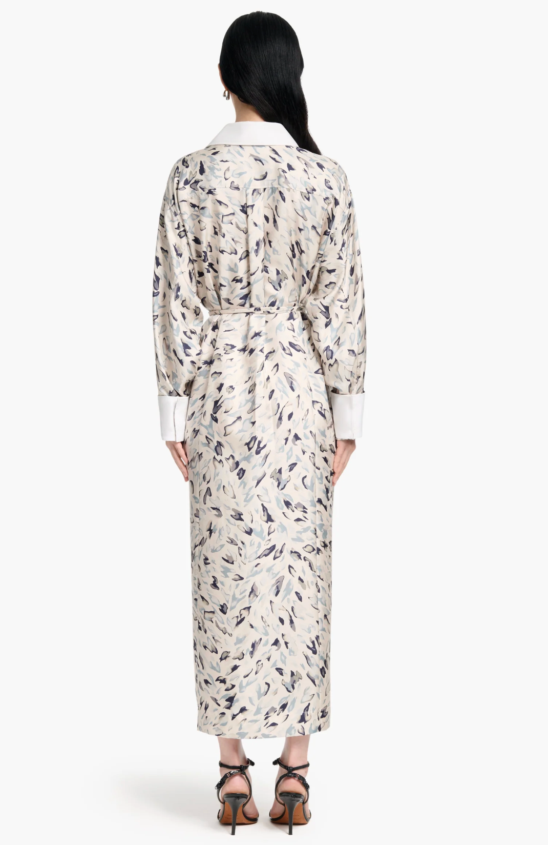 A person stands facing away, wearing the Altuzarra Floyd Dress with a shirt silhouette, light color, white cuffs and collar, paired with black heeled sandals.