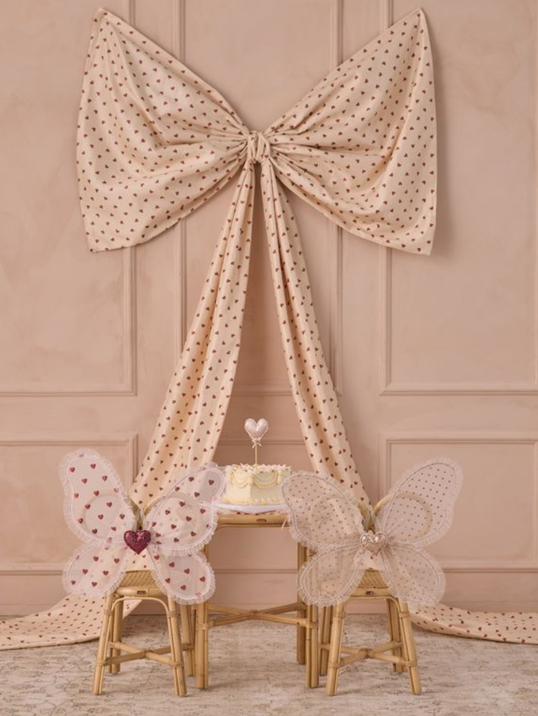 The Noralee Girls' Fairy Set features two butterfly-wing kids’ chairs, a cake-topped round table, and a large polka-dot bow backdrop, creating the perfect dress-up party scene against a light pink wall.